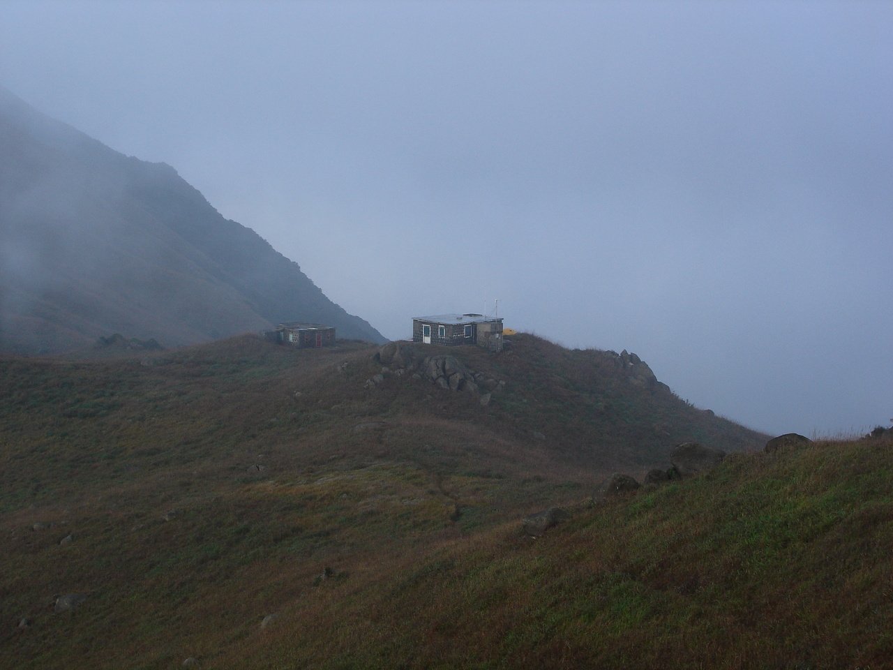 Stone huts on the ridge of Sunset Peak in cloud, with the trail running across the grassland